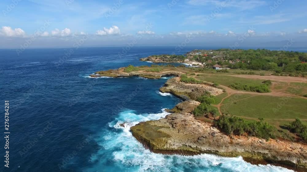 Flying over rocky coastline covered with grass and paths towards beach and village. Foamy waves of turquoise Indian Ocean crash against the rocks on bright sunny day. Aerial shot of Nusa Lambongan. Flying over rocky coastline covered with grass and paths towards beach and village. Foamy waves of turquoise Indian Ocean crash against the rocks on bright sunny day. Aerial shot of Nusa Lambongan.