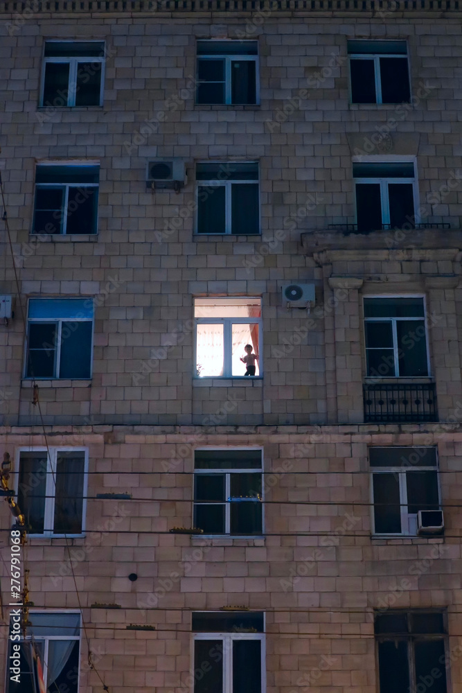 silhouette of a child in the illuminated window of an apartment building in the evening