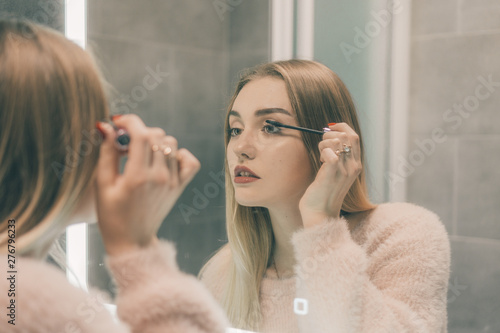 The girl in the bathroom in front of the mirror paints the eyelashes.