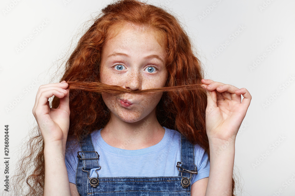 Close up of joyful cute little girl with ginger hair and freckles, looks at  the camera, makes a mustache from strands of hair and looks funny, stands  over white background. Stock Photo |