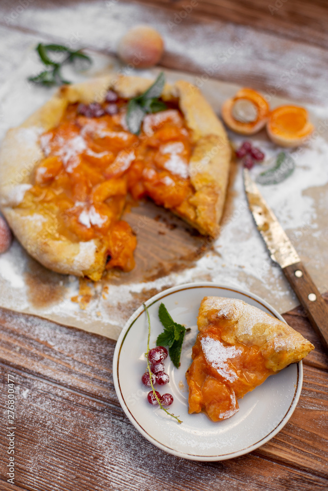 Homemade fruit pie (galette) made with fresh organic apricotes with powdered sugar on wooden table, top view. Plate with a cut piece of cake. Open pie, apricot tart on parchment paper.