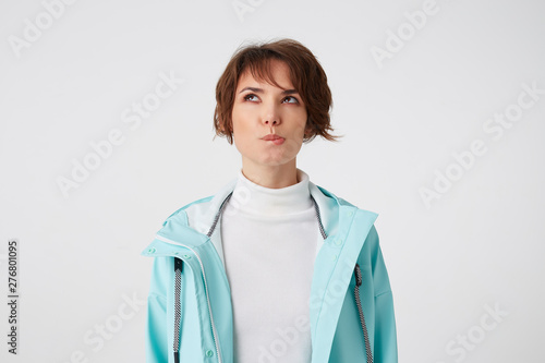 Tablou pe pânză Photo of young wishful short-haired lady in white golf and light blue rain coat, bites lips and looks up, stands over white background