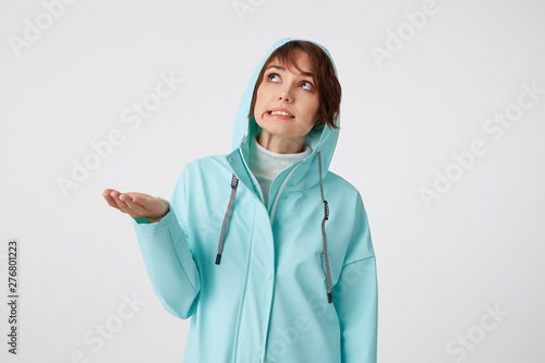 Cute frowning short-haired curly woman in blue rain coat, hiding under the hood from rain and looking up at the rigt side, putting the palm under the rain standing over white background. photo