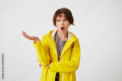 Shocked short-haired young woman in yellow rain coat, with wide open mouth and outraged expression, putting the palm up, frowning and looking away with wide open mouth, stands over white background.