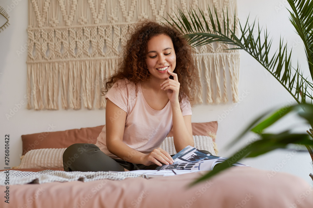 Young african american woman with curly hair, sits on the bed and touches cheek, smiles and reads a new magazine, enjoy free time at home. Young african american woman with curly hair, sits on the bed and touches cheek, smiles and reads a new magazine, enjoy free time at home.