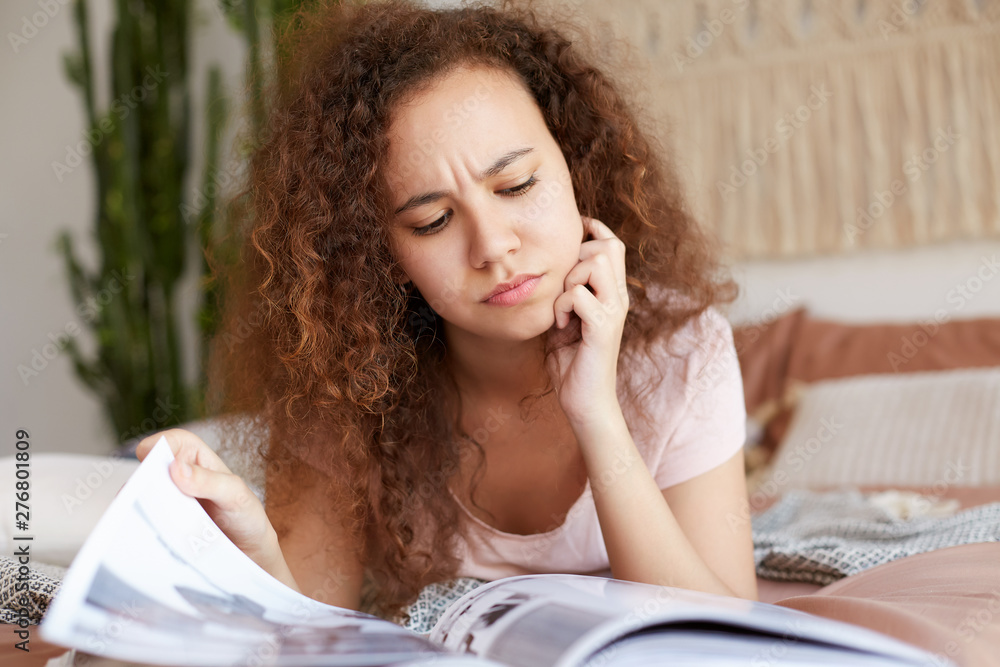Photo of young frowning african american african american lady with curly hair, lies on the bed and touches chin, pensively reading new issue of favorite magazine. Photo of young frowning african american african american lady with curly hair, lies on the bed and touches chin, pensively reading new issue of favorite magazine.