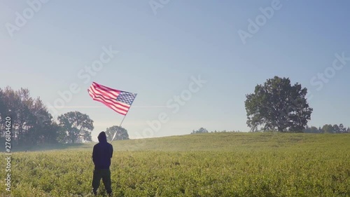 Man, silhouetted, lit by sun, stands on boulder, holds American flag that waves. photo