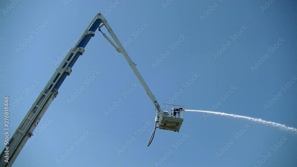 Firefighter from a lifting tower directs a narrow stream of water from a fire pump - water cannon. Side and bottom view. Blue sky background. Firefighter from a lifting tower directs a narrow stream of water from a fire pump - water cannon. Side and bottom view. Blue sky background.