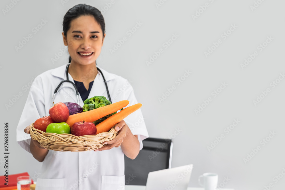 A young woman doctor nutritionist smiling siting working on her laptop in her office holding a basket of vegetables. A young woman doctor nutritionist smiling siting working on her laptop in her office holding a basket of vegetables.