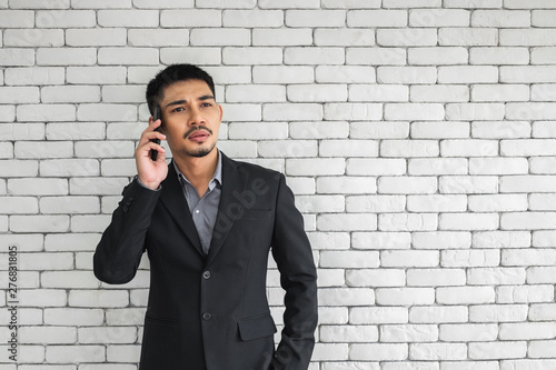 An Asian man in business suit on mobile phone and looking concern, standing in front of white brick wall.