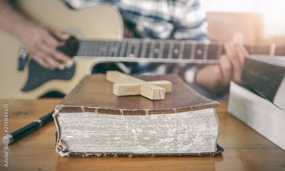 Close up the old Bible and wooden cross with young male playing guitar acoustic at background. Christian worship God concept.
