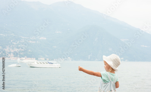 little girl in summer dress and hat looks at the ships on the sea and points his finger at him