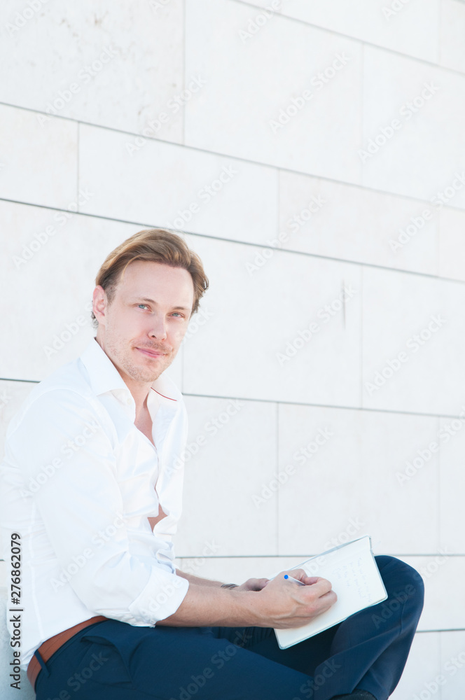Portrait of handsome entrepreneur writing in planner. Positive Caucasian businessman sitting outdoors and writing thoughts in journal Portrait of handsome entrepreneur writing in planner. Positive Caucasian businessman sitting outdoors and writing thoughts in journal