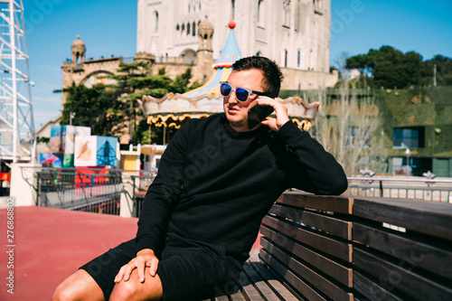 Young handsome man sitting on the bench and talking on the mobile phone on Tibidabo background. Travel concept.