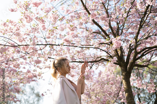 Beautiful blonde young woman in Sakura Cherry Blossom park in Spring enjoying nature and free time during her traveling tourist free time - Wearing white pants and t-shirt with a beige jacket