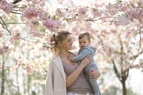 Young mother mom holding her little baby son boy child under blossoming SAKURA Cherry trees with falling pink petals and beautiful flowers