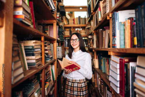 people, knowledge, education and school concept - happy student girl or young woman with book posing in an old library