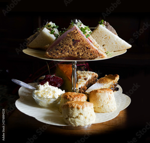Afternoon tea food – sandwiches, scones, cake, carrot cake, apple pie, jam, clotted cream – on a cake stand against a black background in raking light. The skimming light gives it a glamorous touch