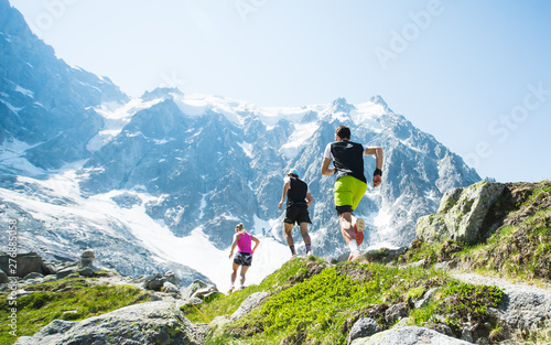 A group of trail runners running along a high mountain trail in the Alps under a clear blue sky.