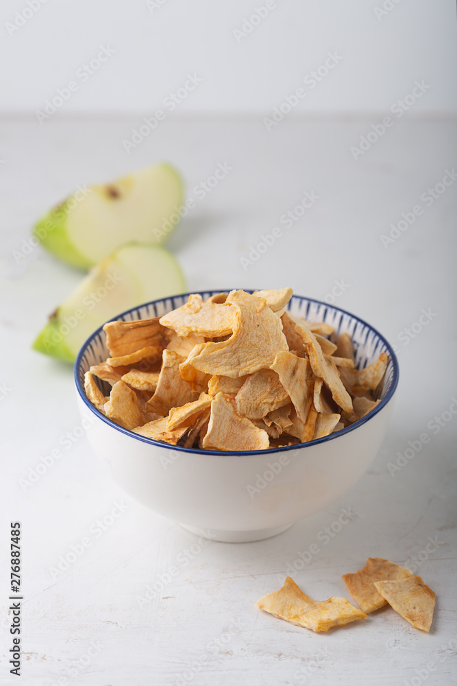 Apple chips in a white bowl and pieces of this fruits on a table