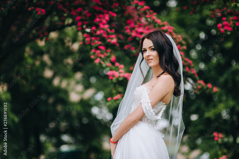 A beautiful and cute bride. A brunette bride with curly hair is standing in a garden with blooming pink flowers. Wedding portrait of a gorgeous model. Concept and photography.