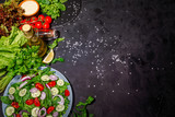 Various fresh mix salad leaves with tomato and cucumber in a glass bowl on dark background with ingredients. Top view with copy space