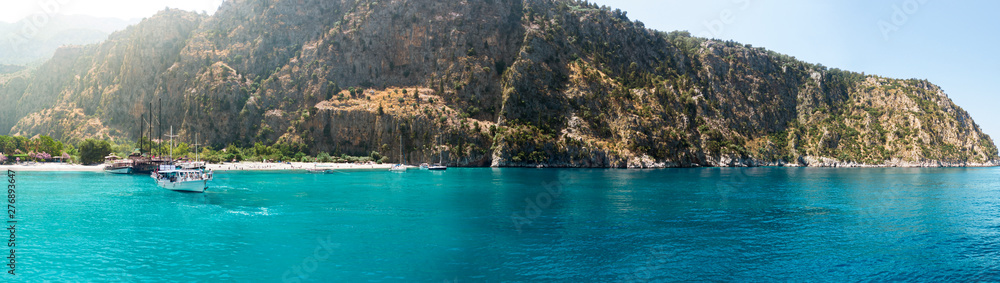 Secluded beach surrounded by valley cliffs in a tranquil bay with turquoise water and sailing boats at sunrise, Oludeniz, Turkey panoramic