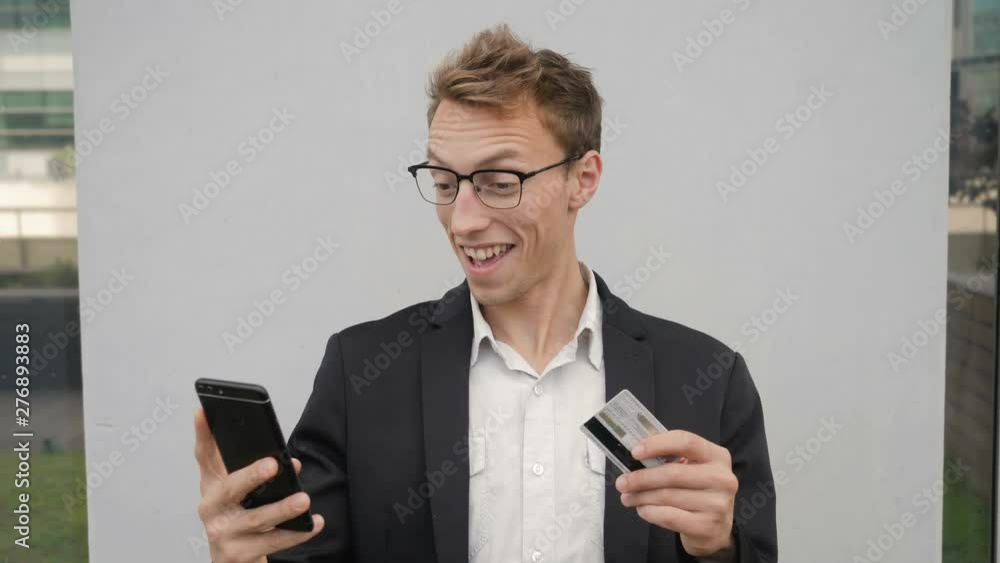 Young Caucasian man in spectacles holding phone and bank card in hands, paying online, finding discount, looking happy and surprised. Lifestyle, online shopping concept Young Caucasian man in spectacles holding phone and bank card in hands, paying online, finding discount, looking happy and surprised. Lifestyle, online shopping concept