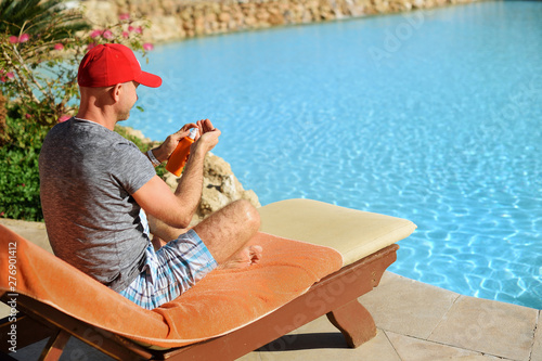 Young and successful man on a sun lounger squeezes out sunblock cream from a tube at the hotel near sweeming pool. Concept summer time to travel