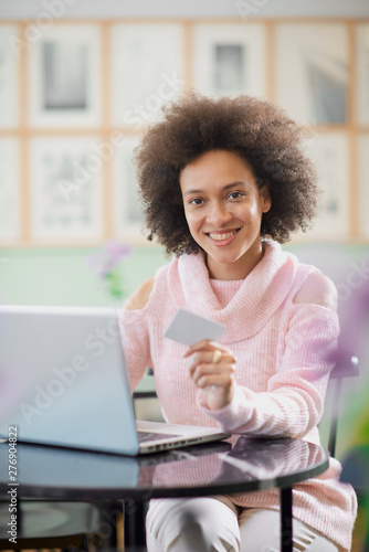 Beautiful mixed race woman in pink turtleneck sweater sitting in pastry shop and using laptop for on line shopping.