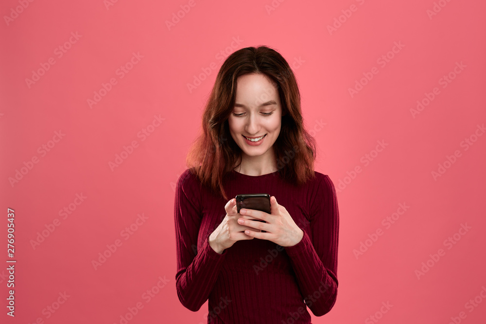Happy pretty brunette girl loooking at the screen of a mobile phone while standing on a dark pink background. Happy pretty brunette girl loooking at the screen of a mobile phone while standing on a dark pink background.