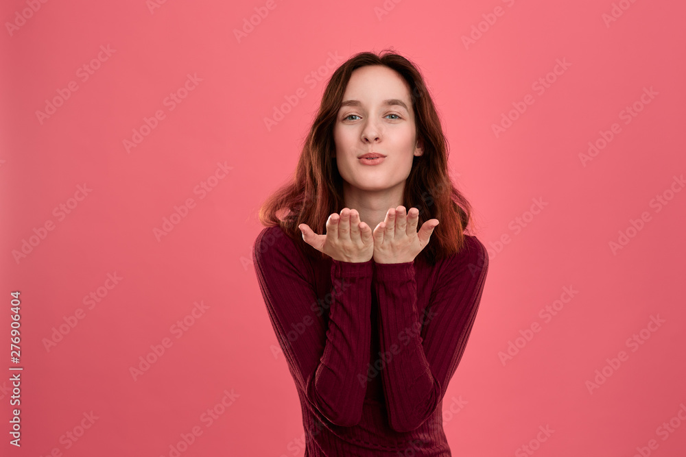 Happy young lady standing isolated over dark pink background while sending romantic hand kisses.