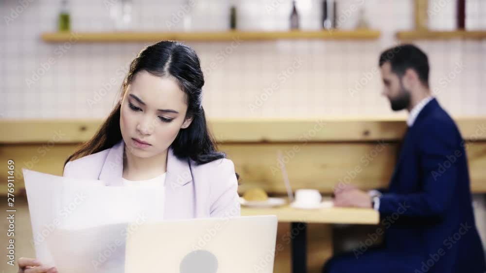 serious businesswoman looking at papers and laptop screen, businessman typing on computer and drinking coffee on background serious businesswoman looking at papers and laptop screen, businessman typing on computer and drinking coffee on background