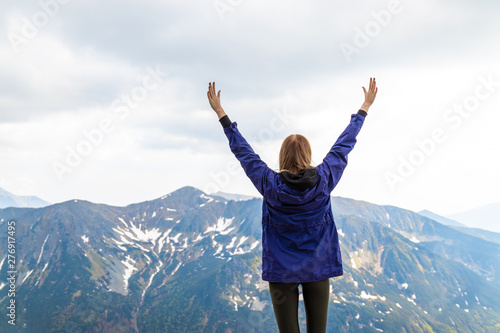 Travel and tourism concept. A young woman feels strong and raised her hands up, enjoys a beautiful mountain landscape on a summer day.