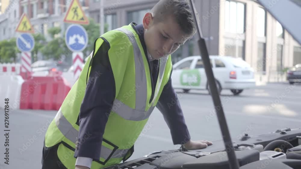 Confident little boy wearing safety equipment standing by the open hood of a broken car. Boy repairing a car. Child as adult. Confident little boy wearing safety equipment standing by the open hood of a broken car. Boy repairing a car. Child as adult.