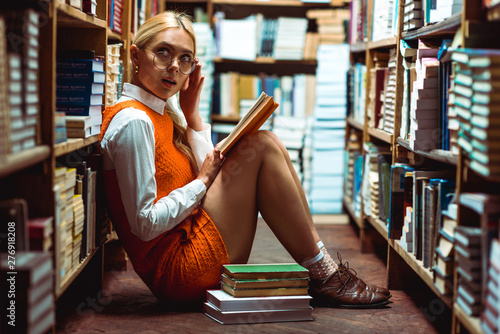 beautiful and blonde woman in glasses holding book and sitting on floor in library