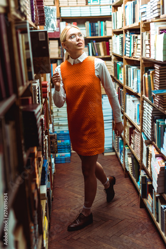 beautiful and blonde woman in glasses and orange dress looking away in library