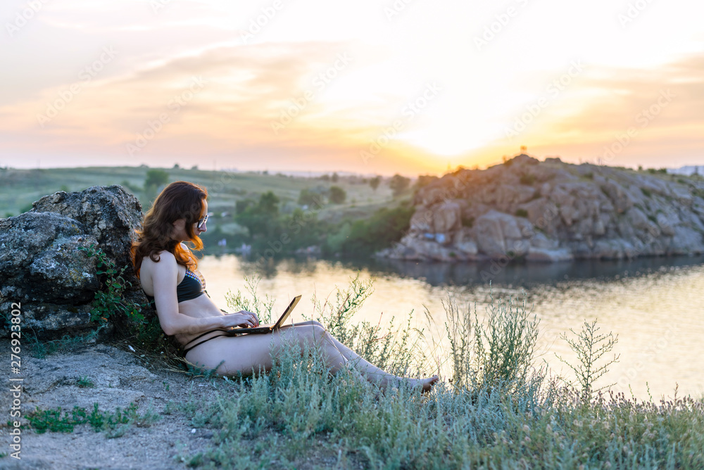 Young freelancer woman working on a laptop while sitting on a rock against the backdrop of a beautiful sunset.