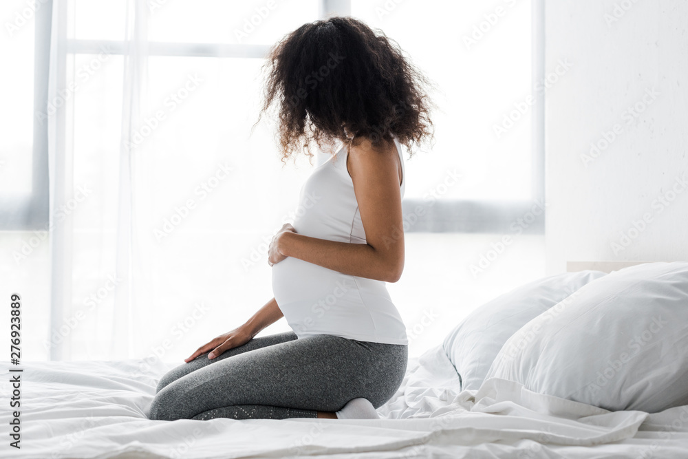 pregnant african american woman touching belly while sitting on bed