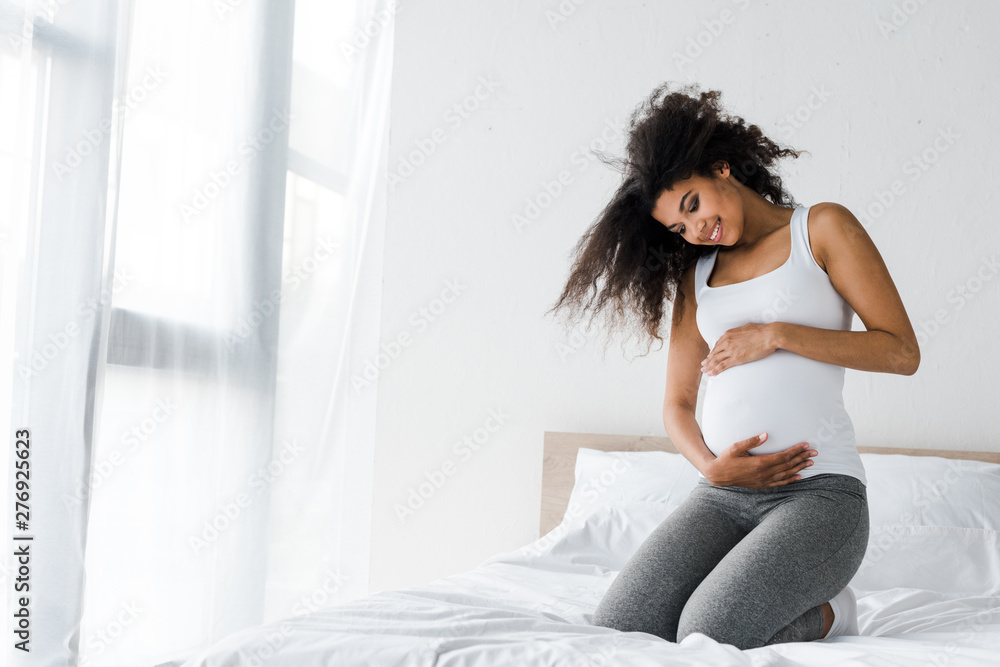 happy pregnant african american woman touching belly while sitting on bed happy pregnant african american woman touching belly while sitting on bed