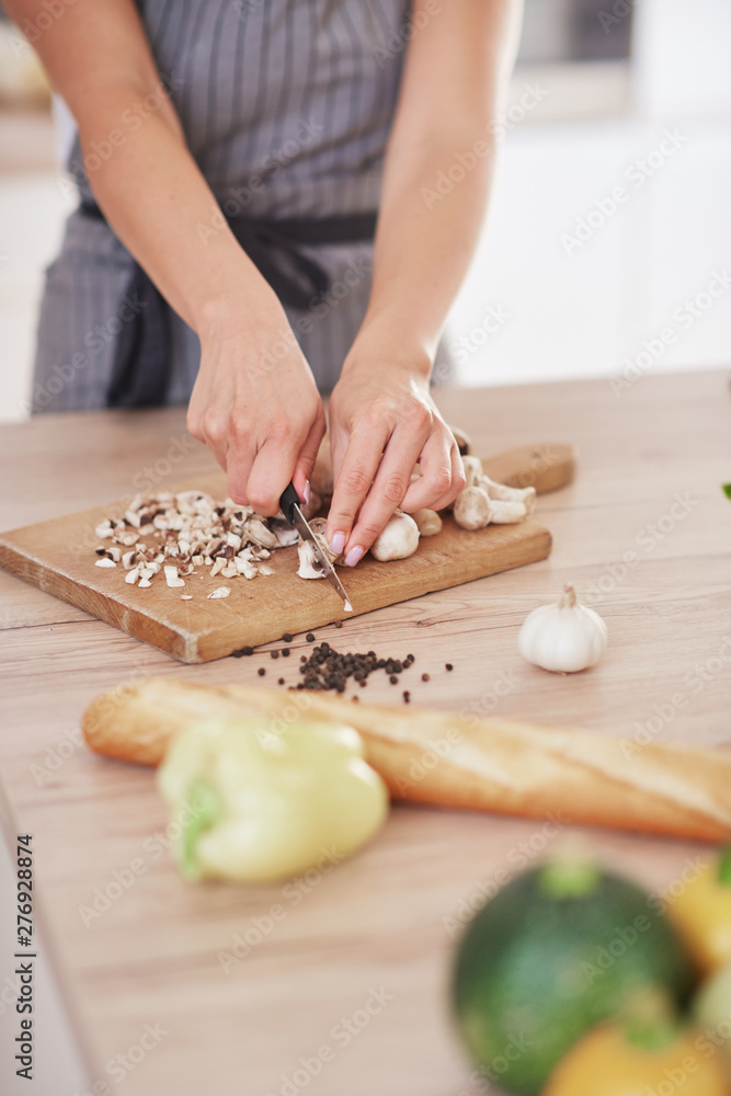 Dedicated Caucasian housewife in apron standing in kitchen and chopping mushrooms. On table are lots of vegetables. Cooking at home concept.