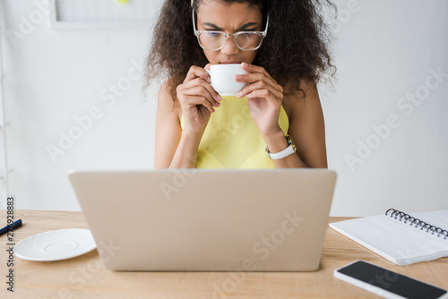 attractive curly african american woman looking at laptop and holding cup