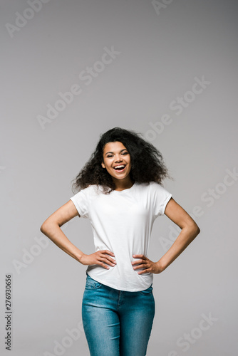 happy young african american woman smiling while standing with hands on hips on grey