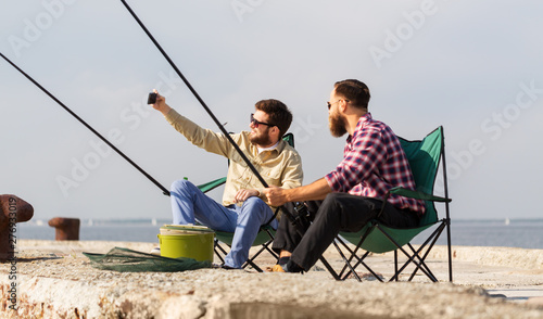 leisure and people concept - male friends with fishing rods taking selfie by smartphone on pier at sea