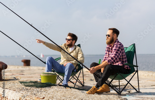 leisure and people concept - male friends with fishing rods sitting in camp-chairs on sea pier