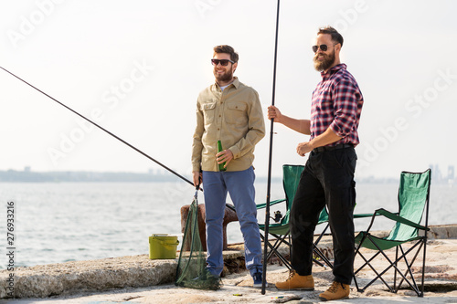leisure and people concept - happy male friends with fishing rods and beer on pier at sea