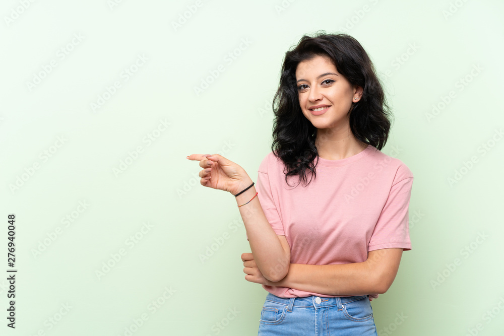 Young woman over isolated green background pointing finger to the side Young woman over isolated green background pointing finger to the side