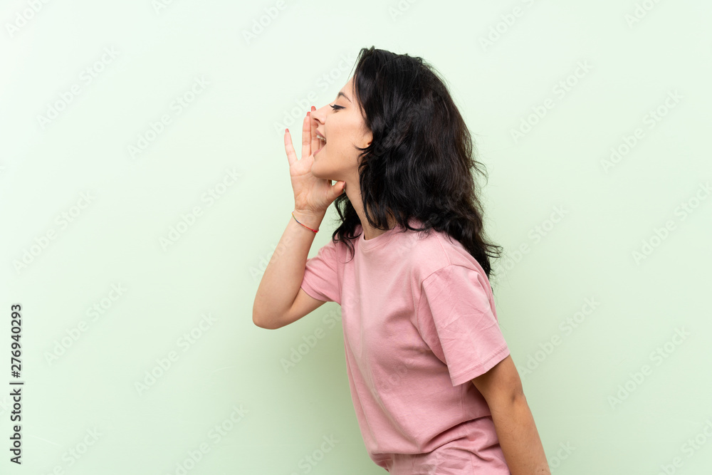 Young woman over isolated green background shouting with mouth wide open to the lateral Young woman over isolated green background shouting with mouth wide open to the lateral