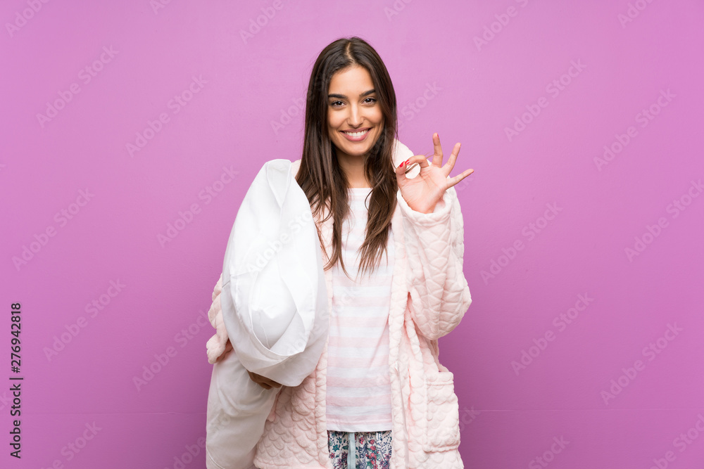 Young woman in pajamas and dressing gown over isolated purple background showing an ok sign with fingers Young woman in pajamas and dressing gown over isolated purple background showing an ok sign with fingers