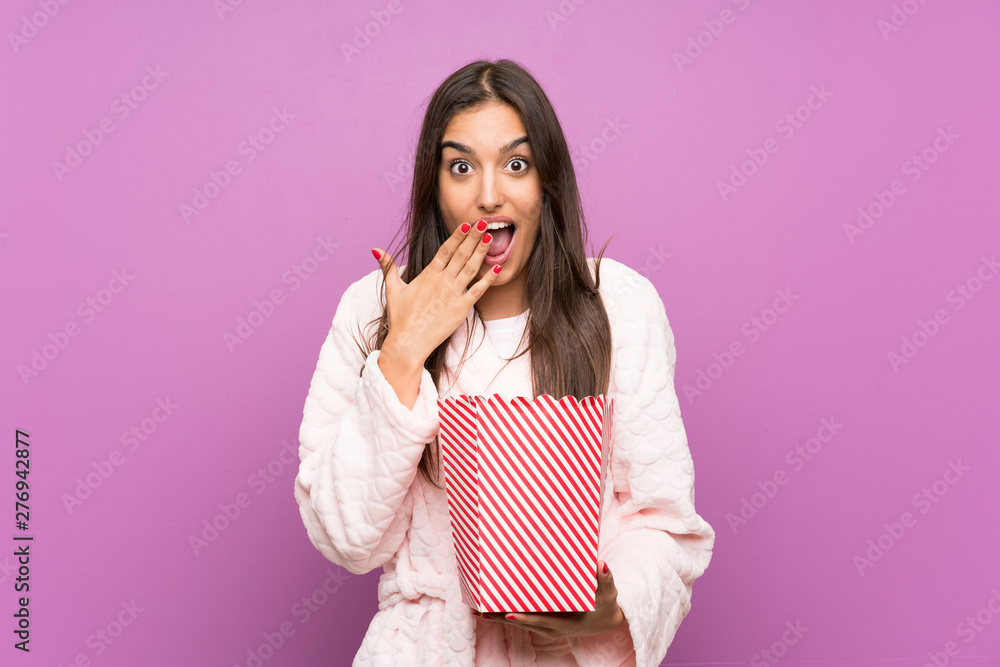 Young woman in pajamas and dressing gown over isolated purple background holding popcorns Young woman in pajamas and dressing gown over isolated purple background holding popcorns